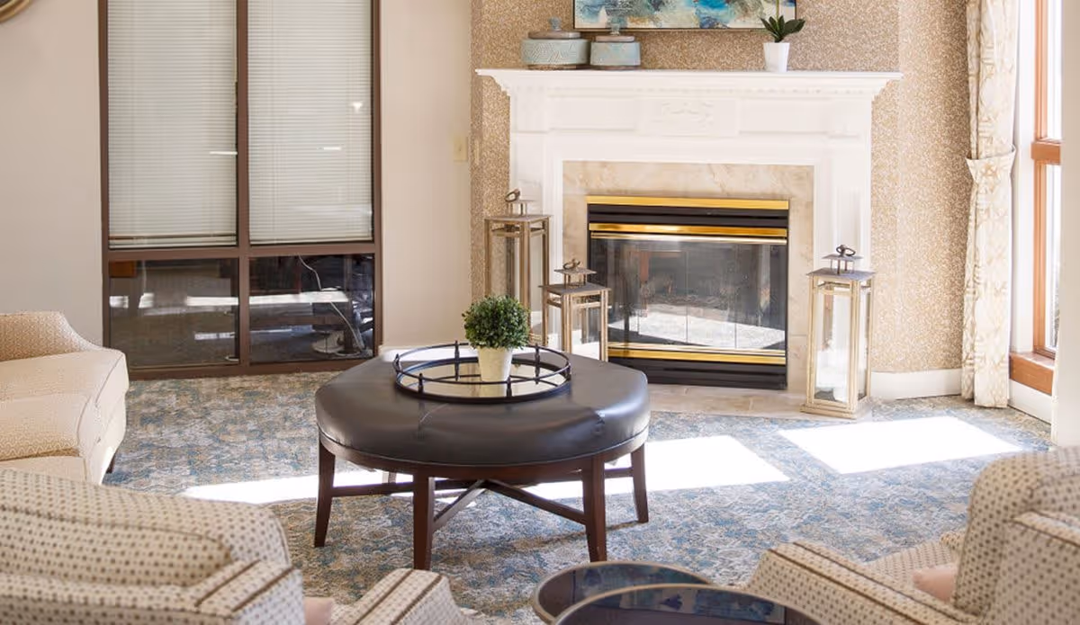 A cozy living room area with a white fireplace in the center, flanked by two large lantern-style candle holders. In front of the fireplace is a round dark leather ottoman with a small potted plant on top. Surrounding the ottoman are patterned armchairs and a beige sofa. The room has a large window with light curtains allowing natural sunlight to brighten the space.