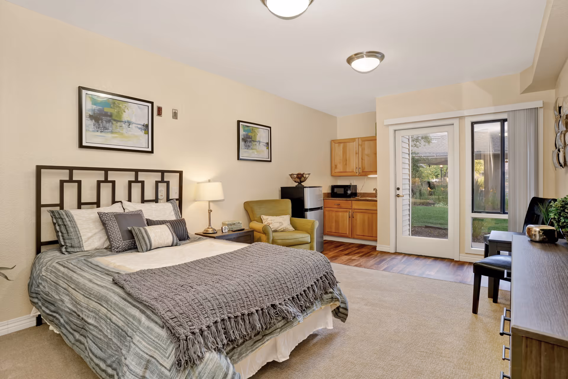 A cozy bedroom in a senior living facility featuring a neatly made bed with multiple pillows and a gray throw blanket. The room includes a green armchair, a small kitchenette with wooden cabinets, a mini fridge, and a microwave. There are two framed abstract paintings on the wall above the bed. A glass door and window provide a view of a garden outside. The room has beige walls and carpeted flooring with a section of wood flooring near the kitchenette.
