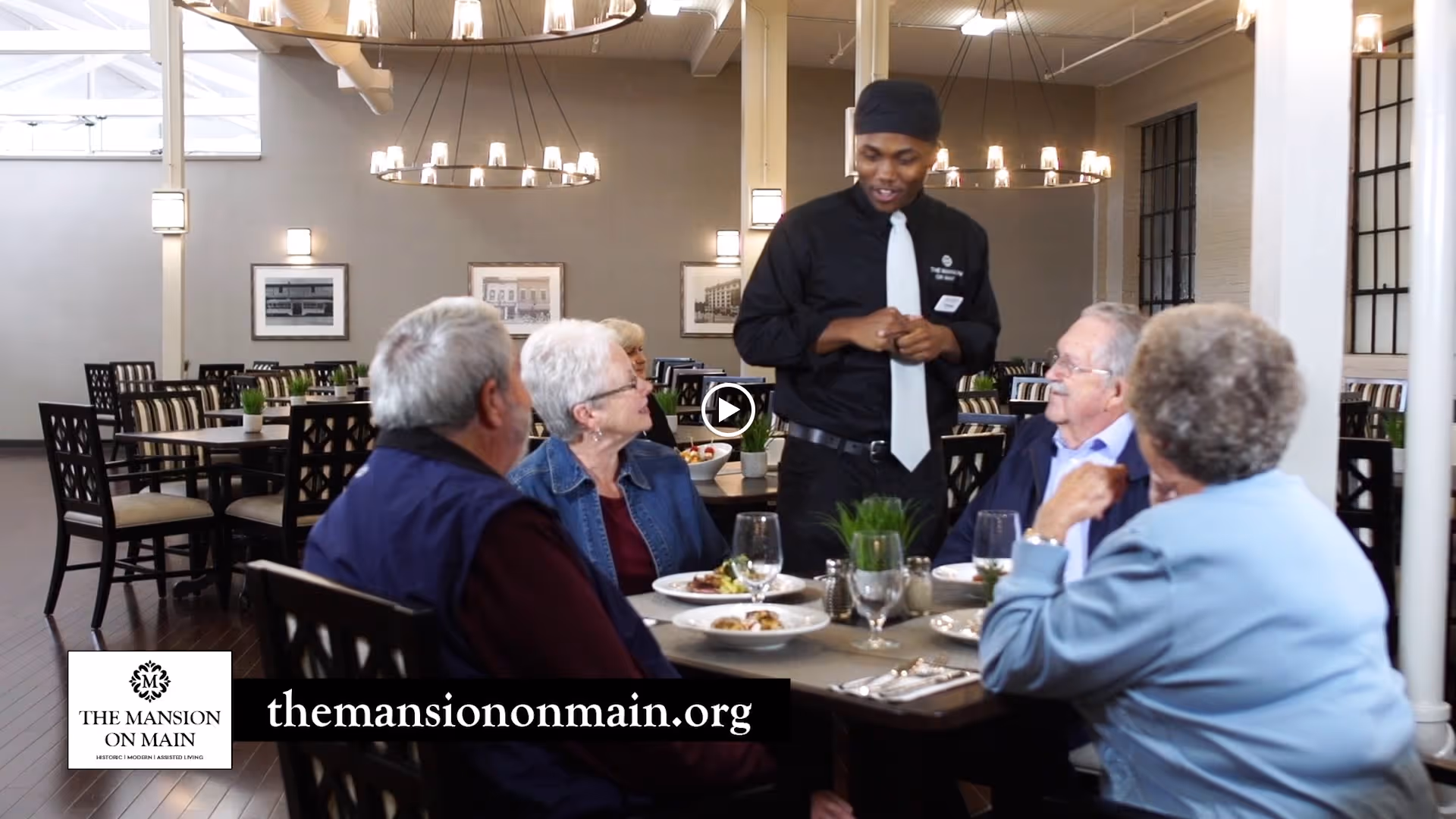 A group of four elderly people sitting around a dining table in a well-lit dining room, being served by a waiter dressed in black with a white tie. The table is set with plates of food, glasses, and a small plant centerpiece. The room has multiple tables and chairs, chandeliers, and framed pictures on the walls.