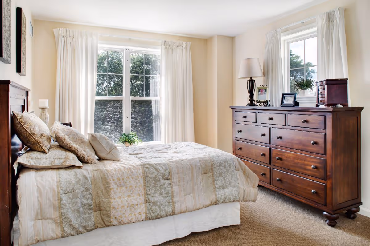 A bright and cozy bedroom with a large bed covered in beige and cream patterned bedding and multiple pillows. There are two large windows with white curtains letting in natural light. A wooden dresser with several drawers stands against the wall, topped with a lamp, framed photos, a small plant, and a decorative box. The walls are painted a soft cream color, and the floor is carpeted.