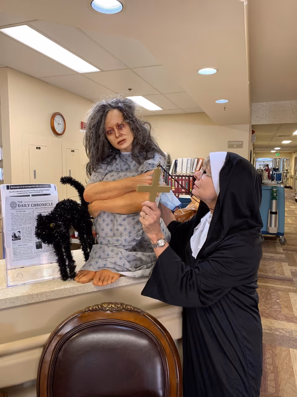 A person dressed as a nun holding a cross up to a life-sized doll with messy hair and dark eye makeup, sitting on a counter next to a black cat decoration and a newspaper stand in a hallway of a senior living facility.