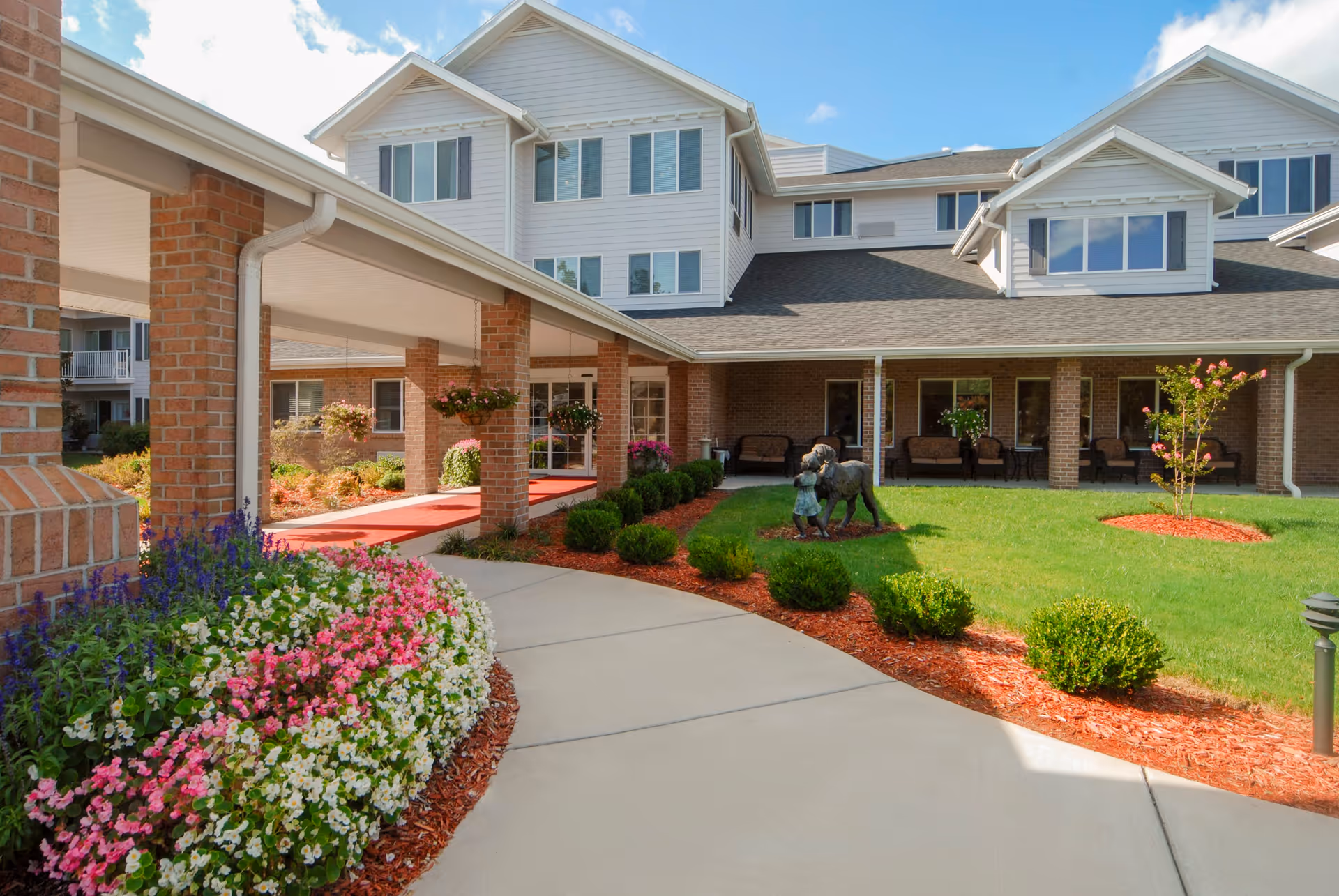 Exterior view of a senior living facility with a covered walkway supported by brick columns, landscaped garden beds with colorful flowers and shrubs, a lawn with a statue of a child and a dog, and a multi-story building with large windows in the background under a partly cloudy sky.