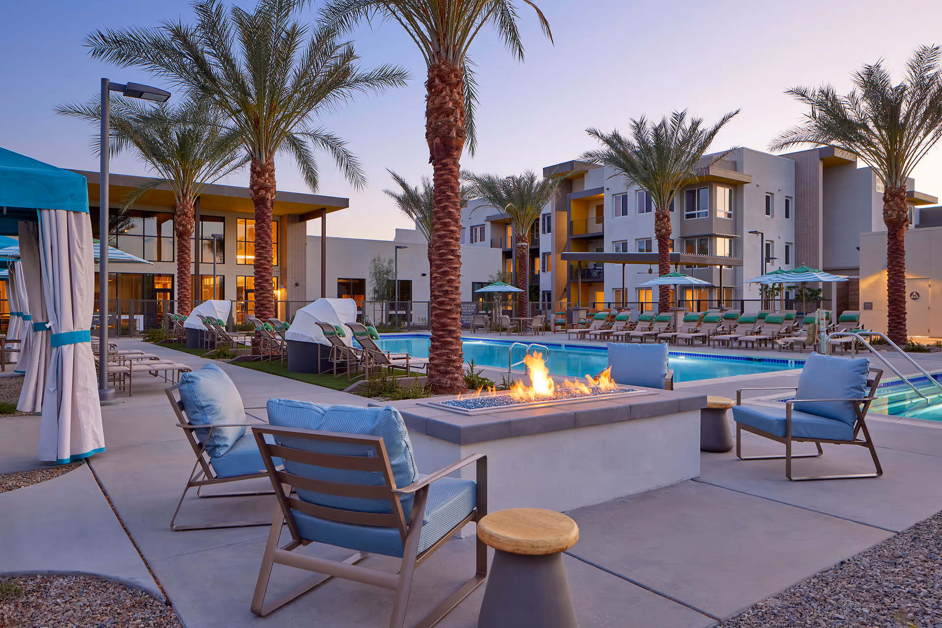 Outdoor seating area with blue cushioned chairs around a rectangular fire pit, palm trees, and a swimming pool with lounge chairs and umbrellas in the background at sunset in a senior living facility.