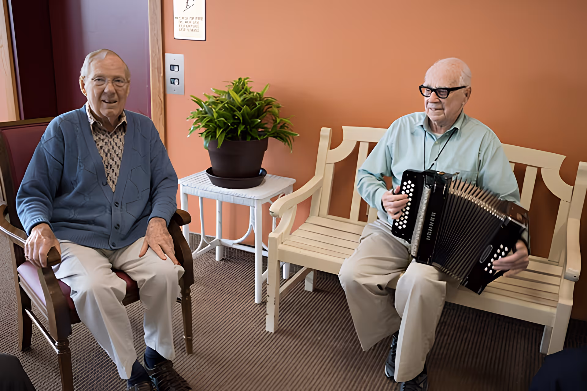 Two elderly men sitting indoors in a room with an orange wall. One man is seated on a wooden chair wearing a blue cardigan and light pants, while the other man is sitting on a white bench playing an accordion. There is a small white table with a potted green plant between them.