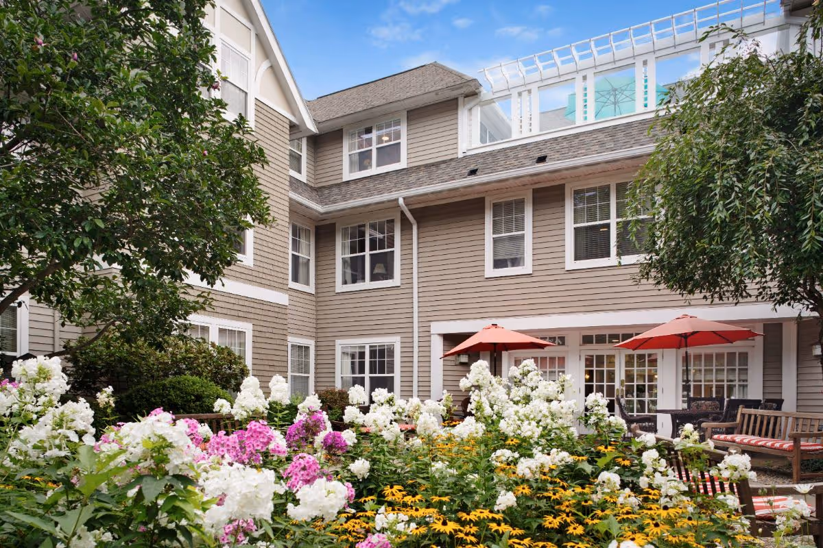Outdoor courtyard area of a senior living facility with blooming white, pink, and yellow flowers in the foreground, surrounded by trees and shrubs. The building has beige siding with white trim and multiple windows. There are wooden benches and tables with red umbrellas for seating.