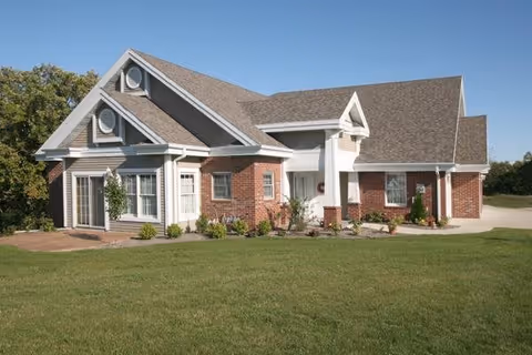 A single-story brick building with white trim and a gabled roof under a clear blue sky, surrounded by a well-maintained lawn and some small shrubs and plants near the foundation.