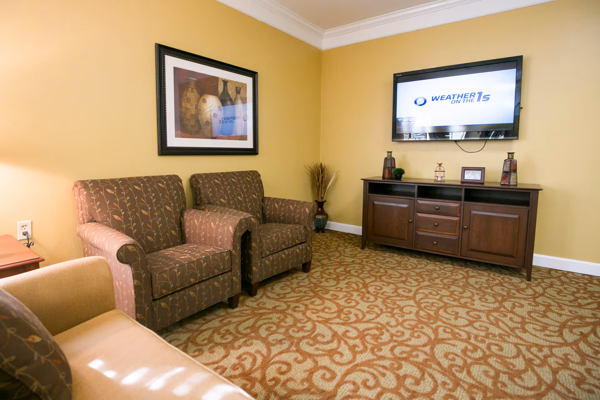 A cozy living room with two patterned armchairs and a beige sofa. A framed artwork hangs on the yellow wall above the armchairs. A flat-screen TV is mounted on the adjacent wall above a dark wooden cabinet with decorative items on top. The floor is covered with a patterned carpet.