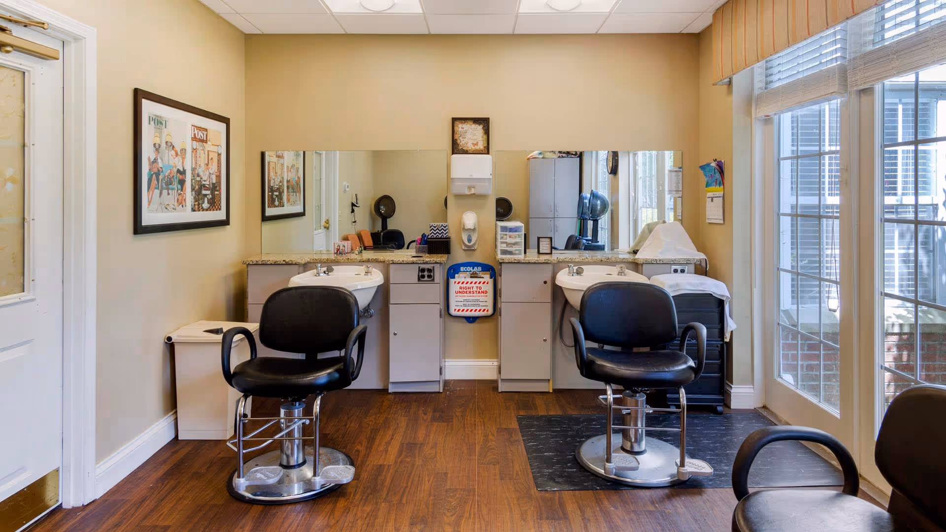 Interior view of a salon area in a senior living facility with two black salon chairs in front of two sinks and mirrors. The room has wooden flooring, beige walls, framed artwork on the wall, and large windows with blinds letting in natural light.