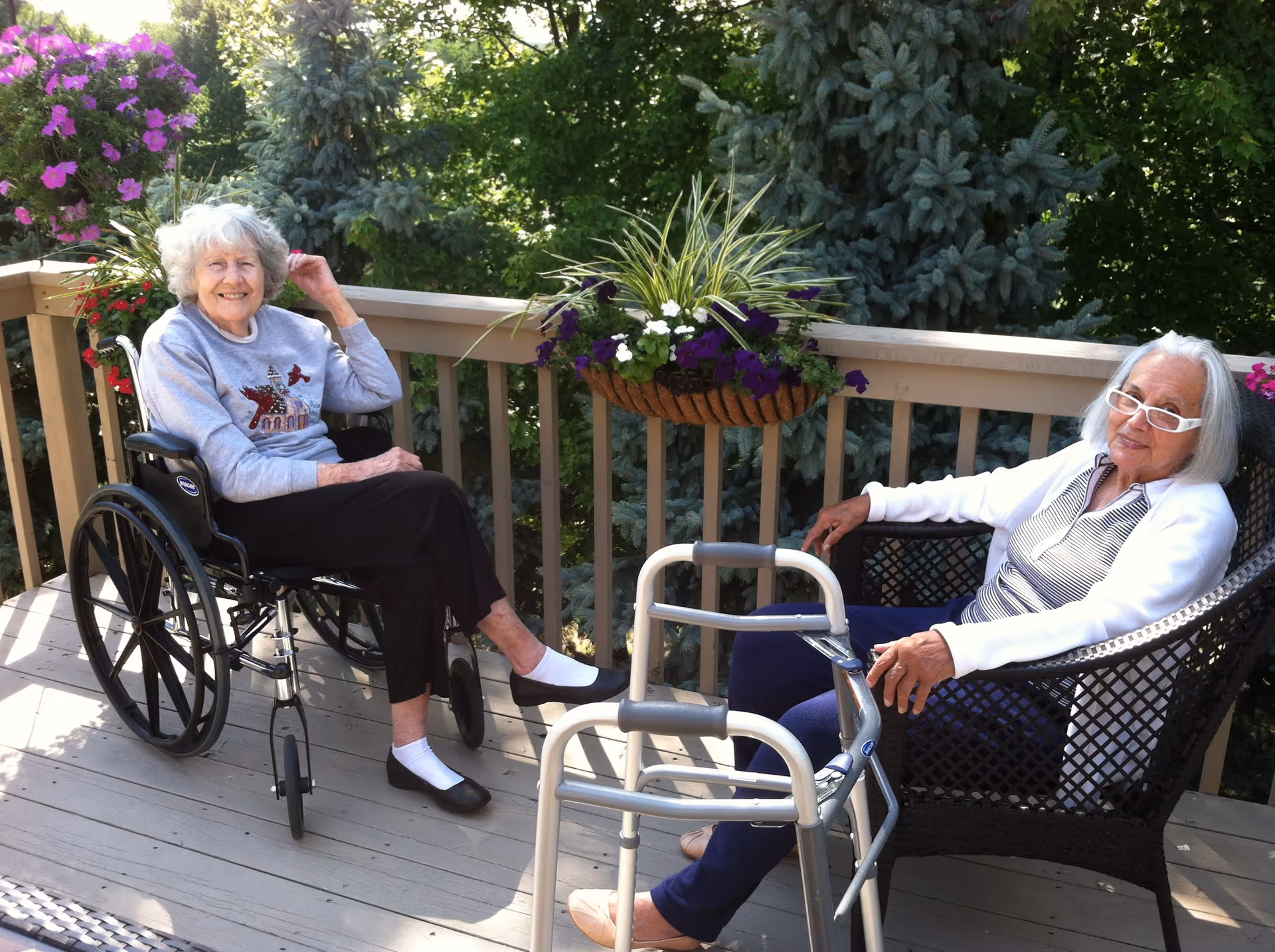 Two elderly women sitting outside on a wooden deck surrounded by greenery and flowers. One woman is in a wheelchair wearing a gray sweater and black pants, while the other woman is seated in a wicker chair wearing a white cardigan and glasses, with a walker in front of her.