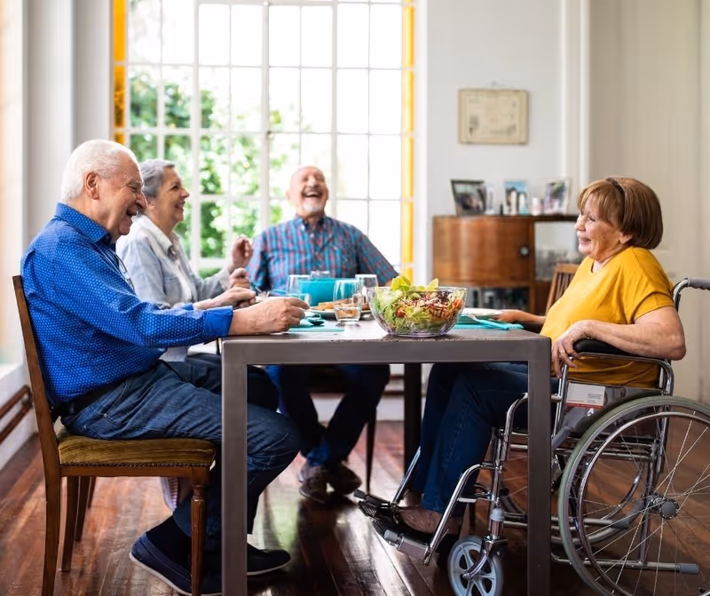 Four elderly people sitting around a dining table in a bright room with large windows. Three of them are seated on chairs, and one woman is in a wheelchair. They are smiling and appear to be enjoying each other's company. There is a bowl of salad and glasses of water on the table.