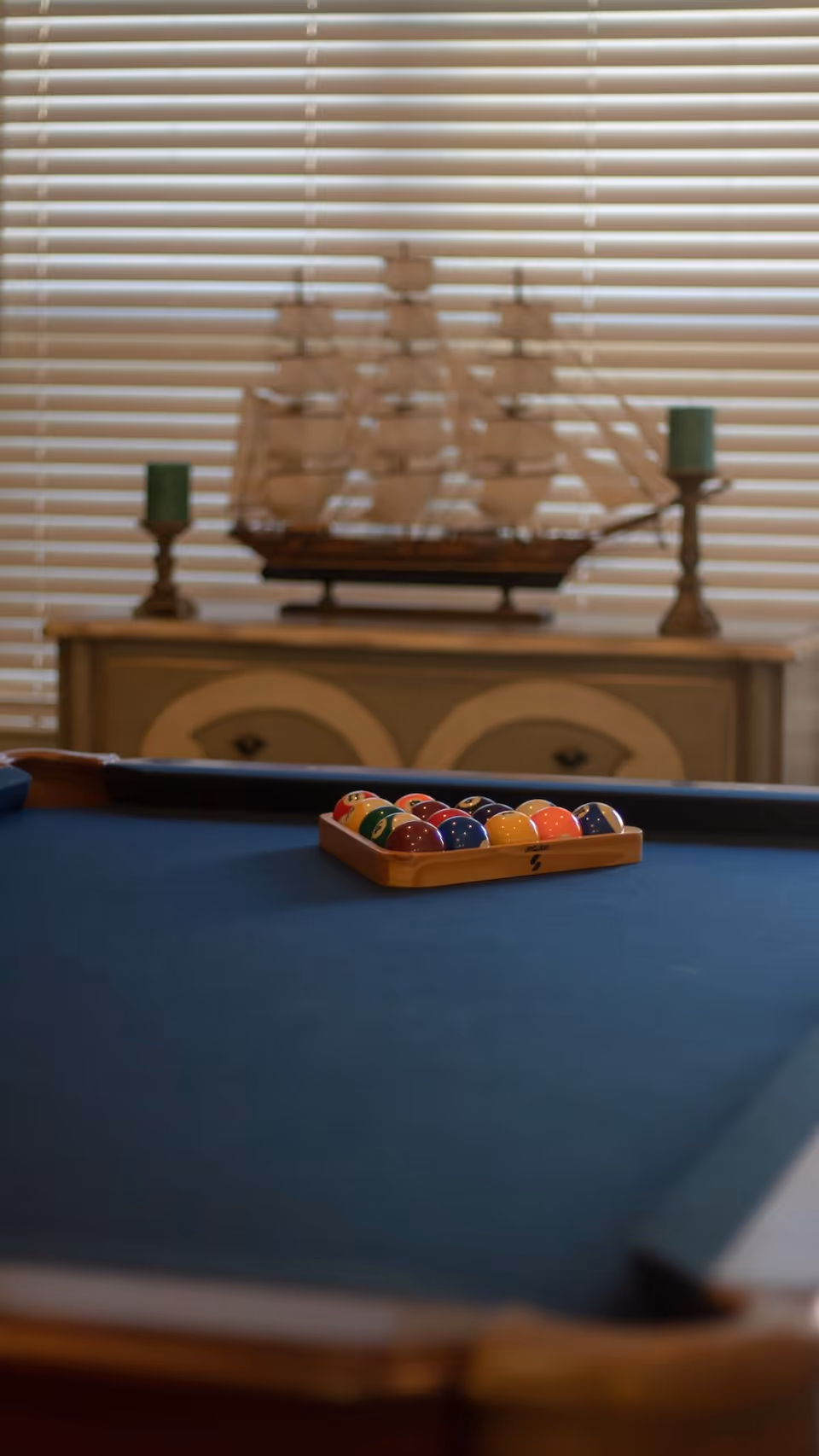 Indoor pool table with a racked set of billiard balls in the foreground and a model ship and candlesticks on a cabinet before closed blinds.