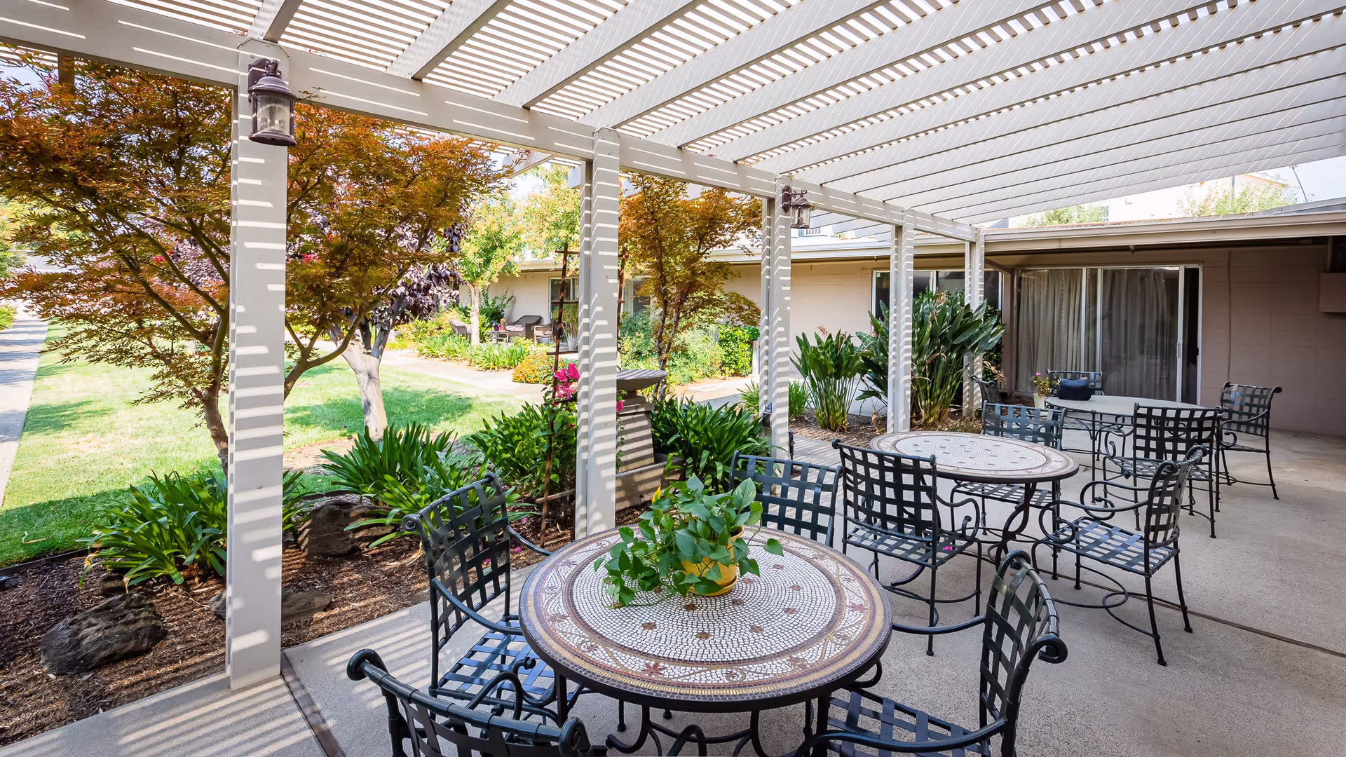 Outdoor patio area with several round mosaic tables and black metal chairs under a white pergola. There are plants and trees surrounding the patio, and a building with sliding glass doors in the background.