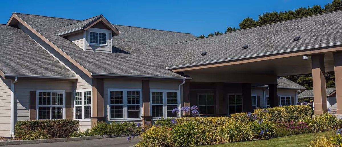 Front exterior of a senior living building with a covered entrance, multiple windows, and landscaped flowerbeds.