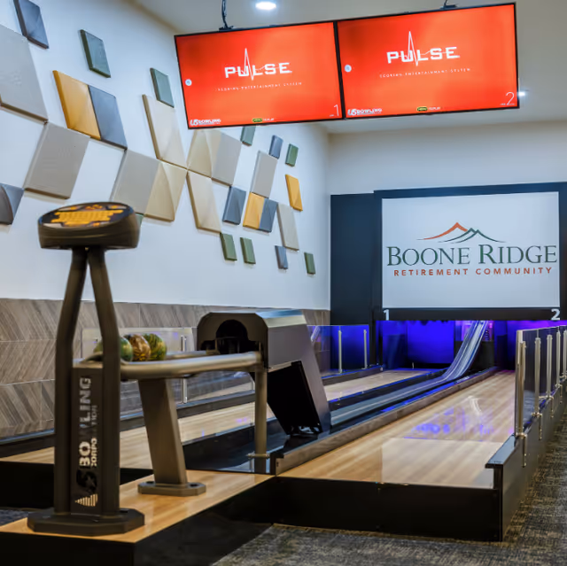 Indoor bowling alley lane with bowling balls on a rack, two large red scoring screens overhead displaying 'PULSE Scoring Entertainment System', and a wall sign that reads 'Boone Ridge Retirement Community'. The wall is decorated with geometric acoustic panels in various colors.