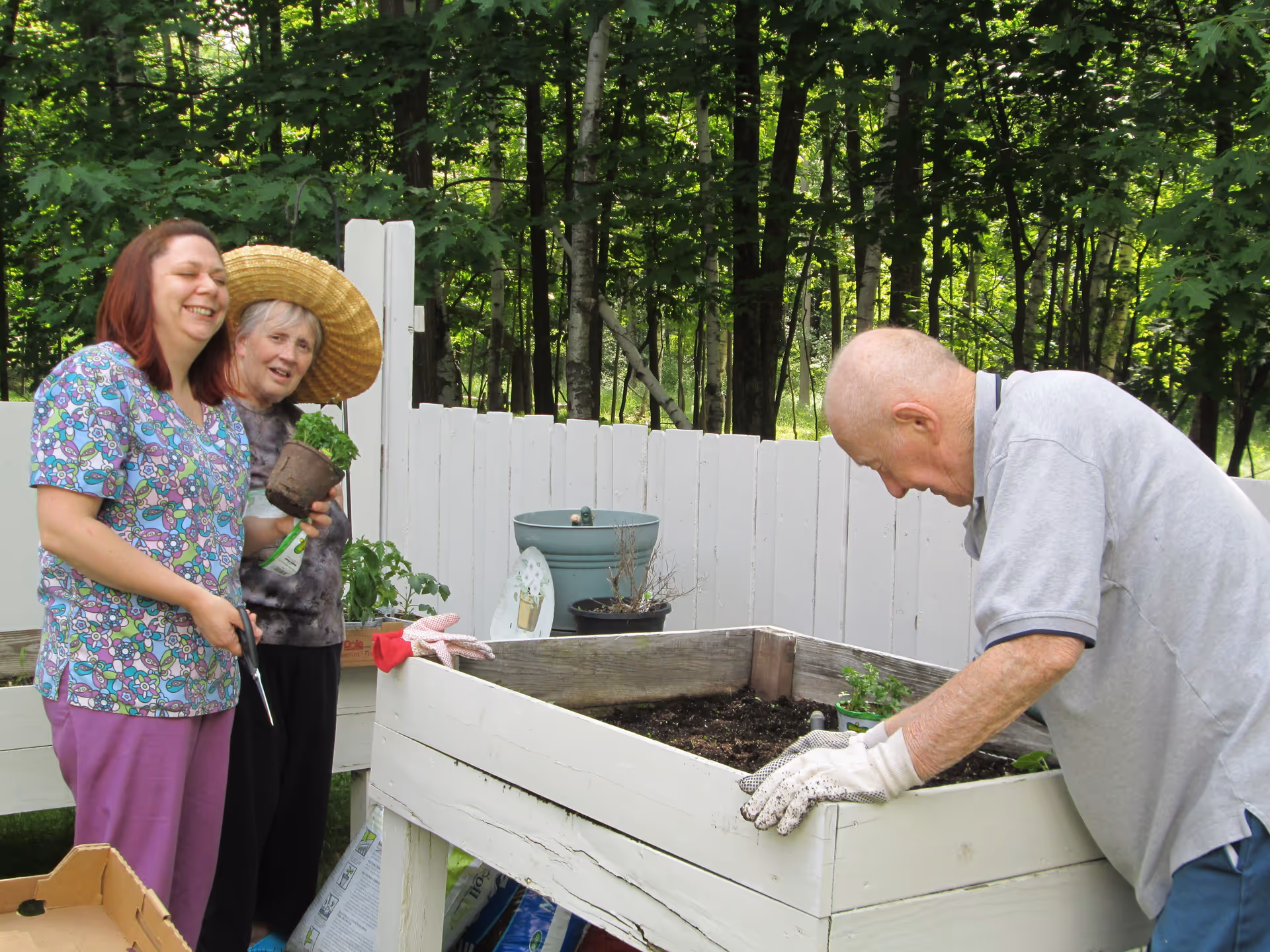 Two women and one man gardening outdoors by a raised white planter box. One woman is holding a small plant and a pair of scissors, smiling, while the other woman wears a large straw hat and stands next to her. The man is leaning over the planter box, wearing gardening gloves and tending to the soil. There is a white wooden fence and green trees in the background.