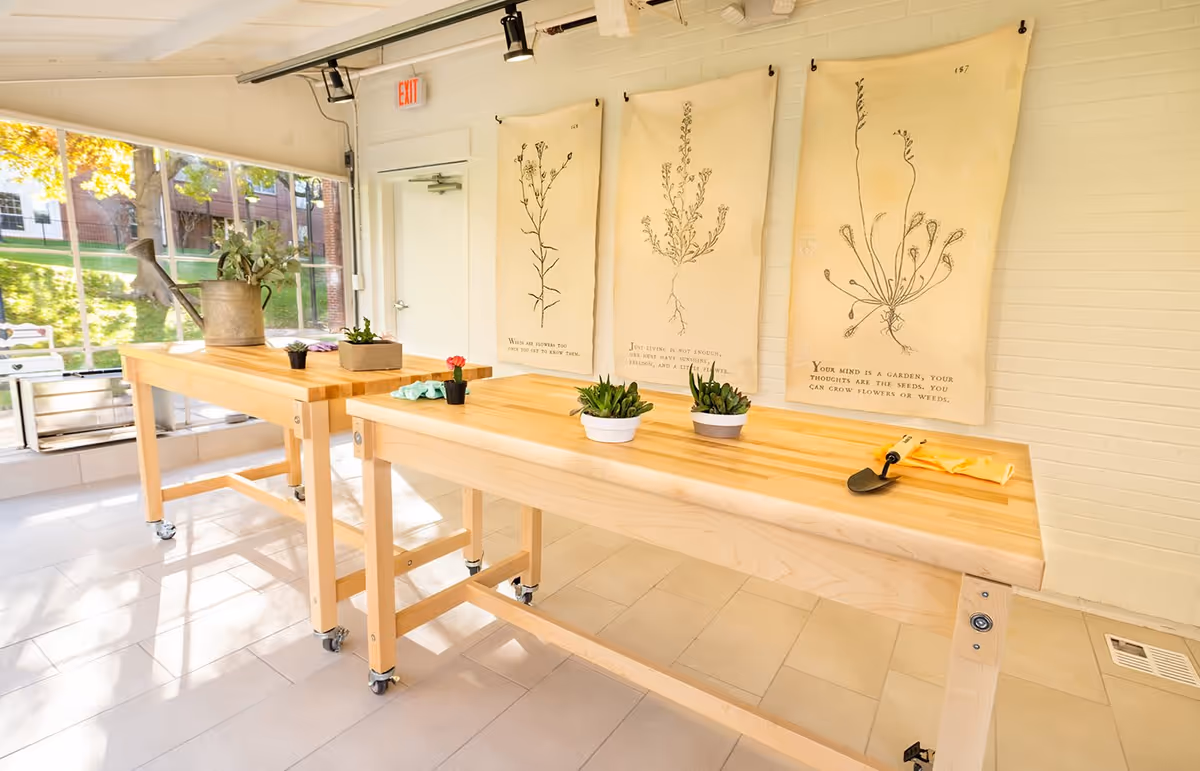 Bright indoor gardening workspace with two wooden tables on wheels, small potted plants, gardening tools, and botanical drawings hanging on a white brick wall. Large windows let in natural light and show green grass and trees outside.
