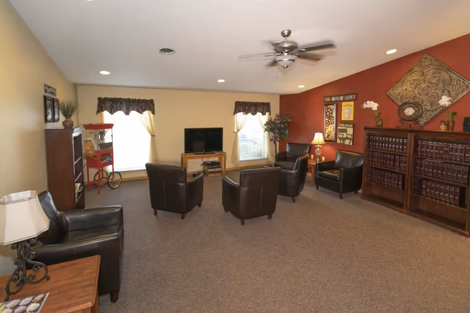 A cozy living room with five dark leather armchairs arranged in a semi-circle facing a flat-screen TV on a wooden stand. The room has beige walls with one accent wall painted red, decorated with framed art and a large ornamental clock. There are two windows with curtains, a wooden bookshelf filled with books, a small table with a lamp, a popcorn machine, and a potted plant in the corner. The ceiling has recessed lighting and a ceiling fan.
