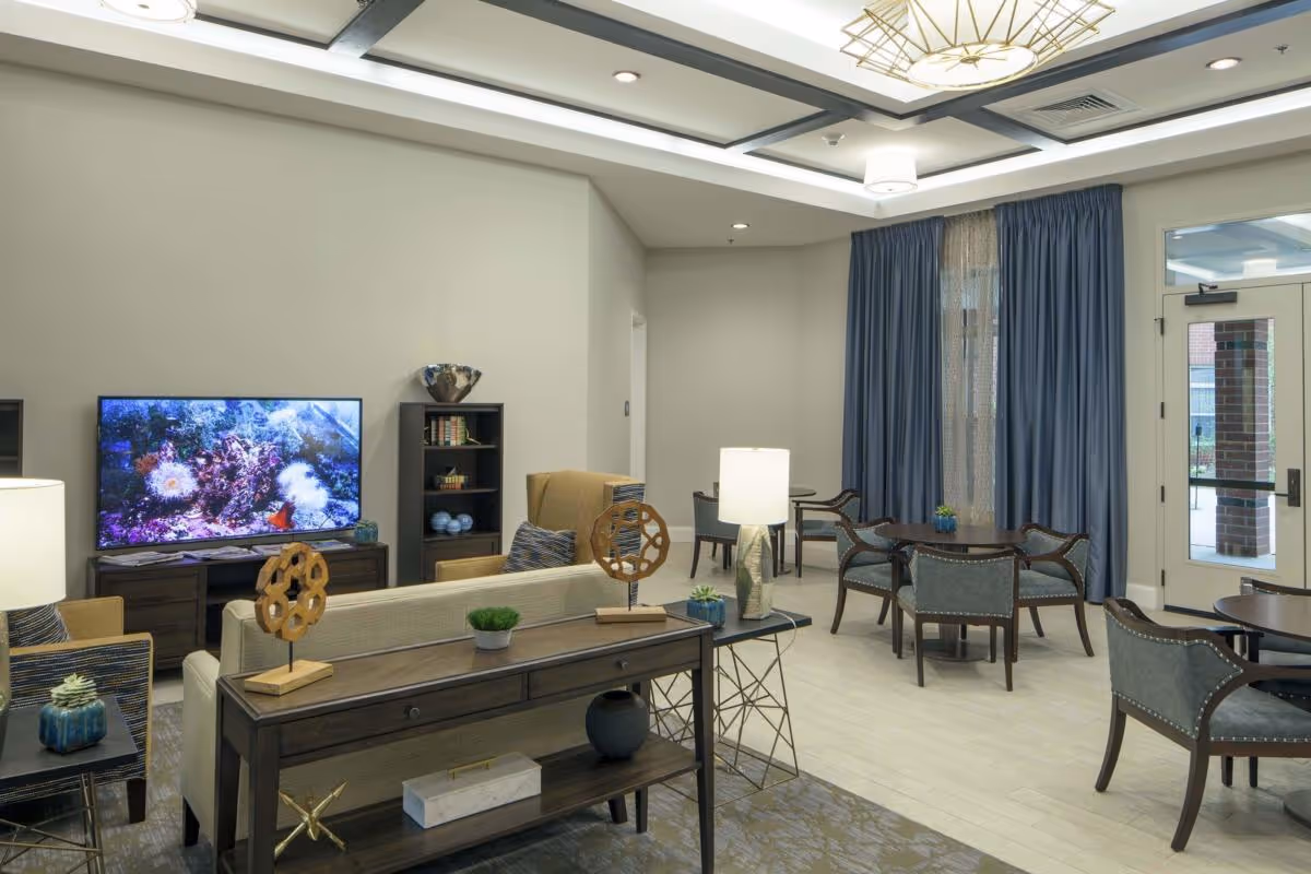 Spacious furnished community living room with sofas, a television, round tables and chairs, and a glass exit door.