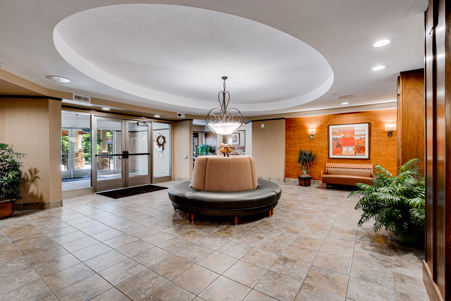 Spacious lobby area with tiled floor, circular cushioned seating in the center, a decorative chandelier hanging from a recessed ceiling, potted plants, a bench against a wooden accent wall with framed artwork, and glass double doors leading outside.