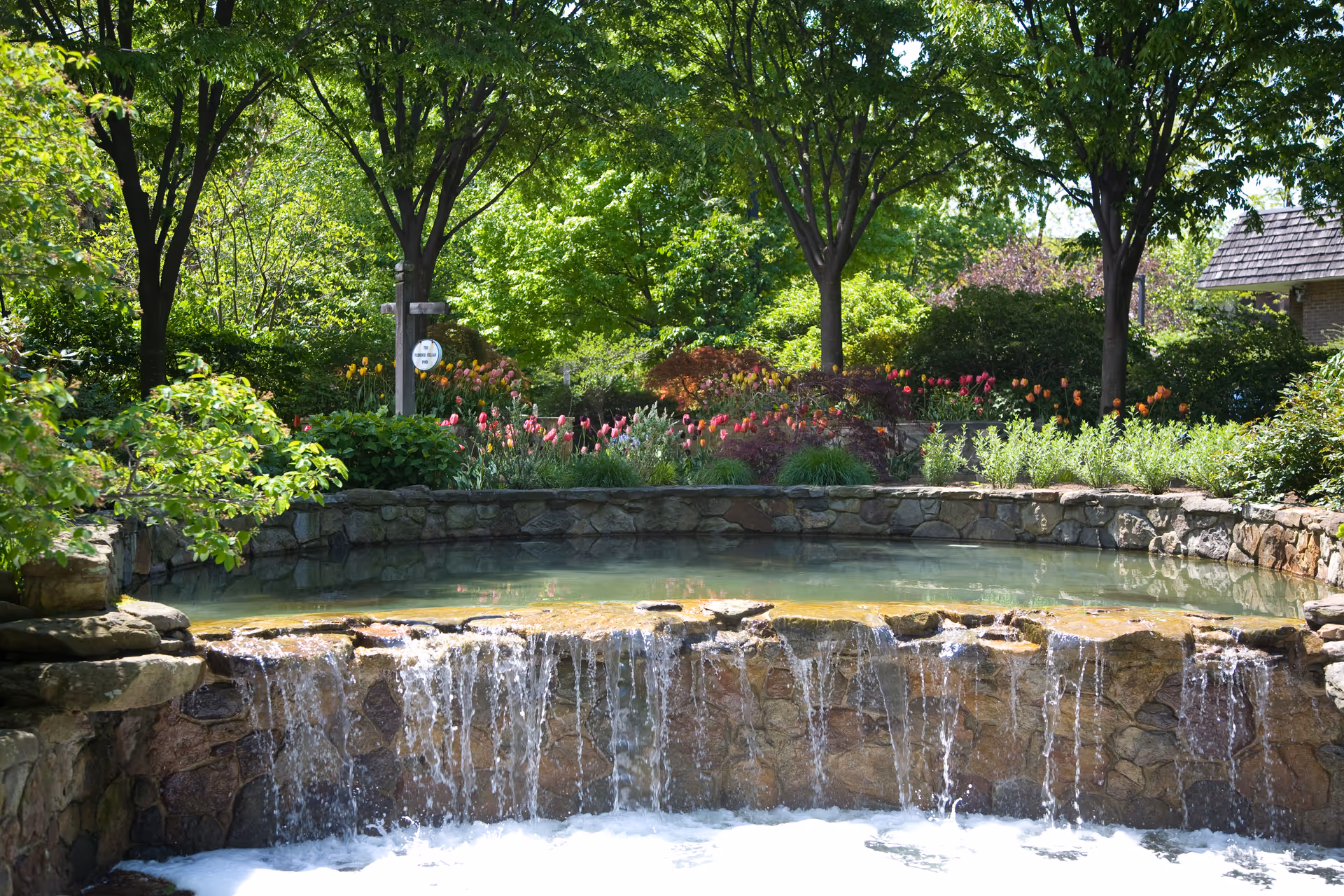A serene outdoor garden area with a stone waterfall feature flowing into a small pool. Surrounding the water feature are lush green trees, colorful tulips, and various shrubs, creating a peaceful and vibrant natural setting.