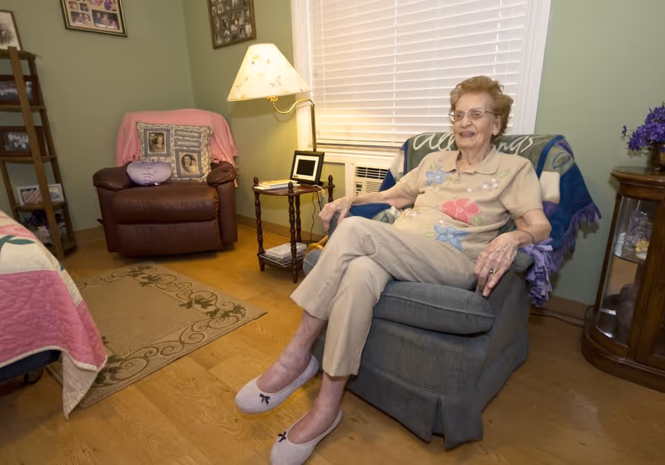 An elderly woman sitting comfortably in a cushioned armchair in a cozy room with wooden flooring. The room has a green wall, a window with blinds, a side table with a lamp, and a brown leather armchair with decorative pillows. There are framed photos on the walls and a small cabinet with flowers on top.