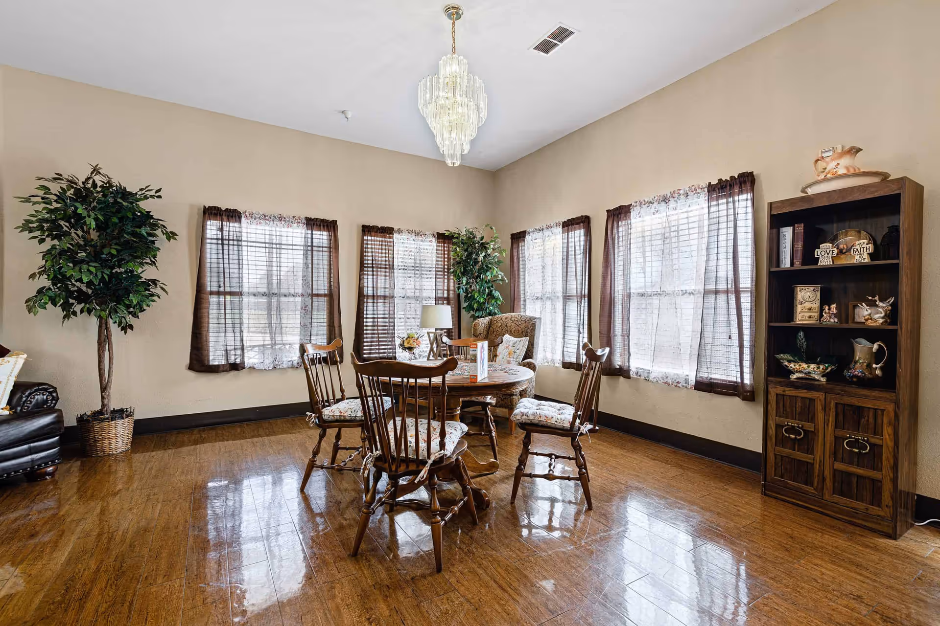 A bright room with wooden flooring and beige walls featuring a round wooden table with four cushioned wooden chairs around it. The room has four windows with sheer white curtains and brown drapes. There is a tall green potted plant in the corner, a wooden bookshelf with decorative items on the right, and a chandelier hanging from the ceiling.