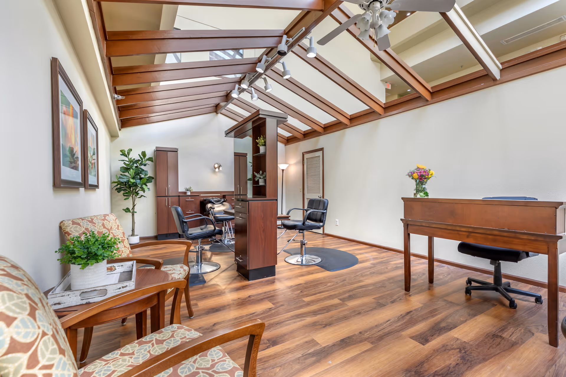 Interior of a bright room with wooden flooring and a slanted glass ceiling with wooden beams. The room contains two patterned armchairs with a small wooden table between them, a tall potted plant, a wooden desk with a black office chair, and a central wooden shelving unit with two black salon chairs on either side. There are framed pictures on the wall and a vase with flowers on the desk.