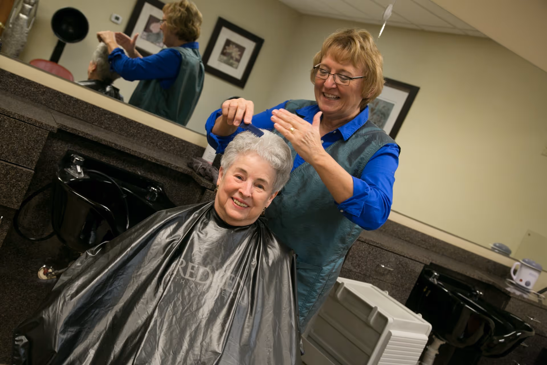 An elderly woman with short gray hair is sitting in a salon chair wearing a black hairdressing cape, smiling as a hairstylist combs her hair. The hairstylist, wearing glasses and a blue shirt with a green vest, is standing behind her. The salon area has black sinks and a large mirror reflecting the scene.