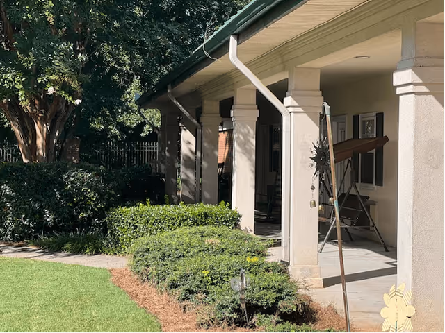 A shaded outdoor patio area with beige columns supporting a roof, surrounded by green bushes and trees. There is a swing on the patio and a well-maintained lawn in the foreground.