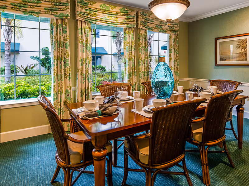 A formal dining room with a wooden table set for six, wicker-style chairs, large windows with floral curtains, and a blue glass centerpiece.