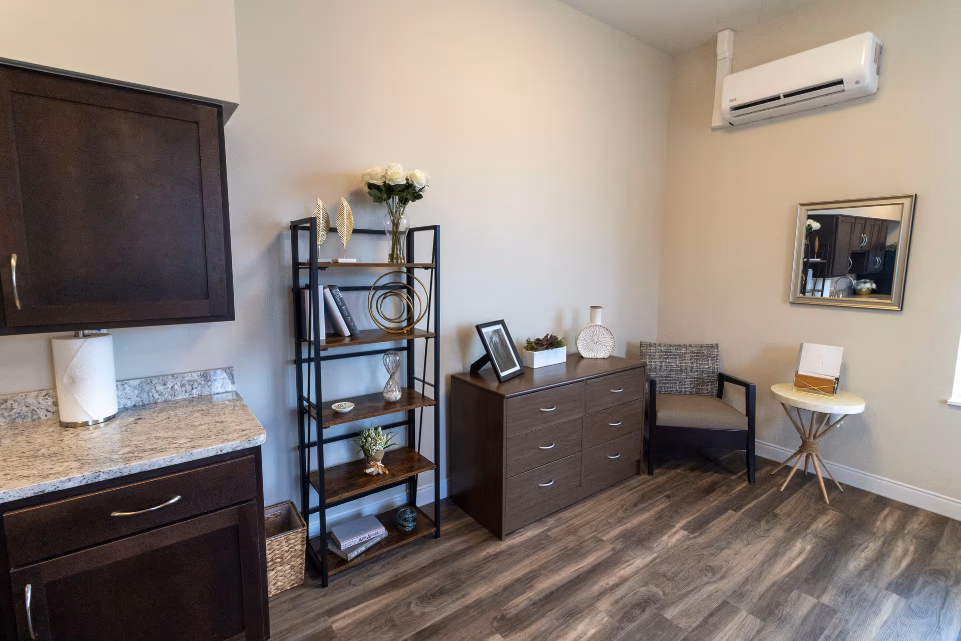 A small furnished sitting area with a bookshelf, dresser, armchair, side table, and kitchenette cabinet.