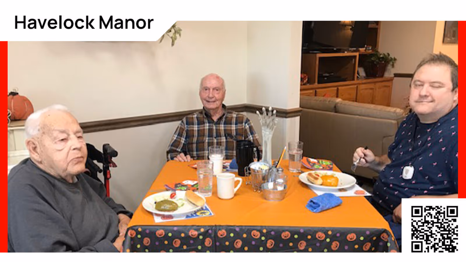 Three men sitting around a table with orange tablecloth decorated with Halloween-themed patterns. They are having a meal with plates of food, cups, and glasses on the table. The room has a cozy interior with a couch and wooden furniture in the background.