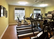A small fitness room with exercise equipment including two treadmills, two stationary bikes, and an elliptical machine. The room has light yellow walls, two windows with blinds, and framed pictures on the left wall.