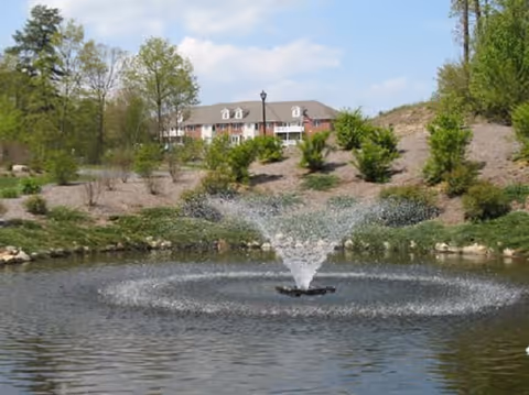 A scenic outdoor view featuring a pond with a water fountain in the center, surrounded by greenery and small trees. In the background, there is a multi-story building partially visible behind the hill and trees under a partly cloudy sky.