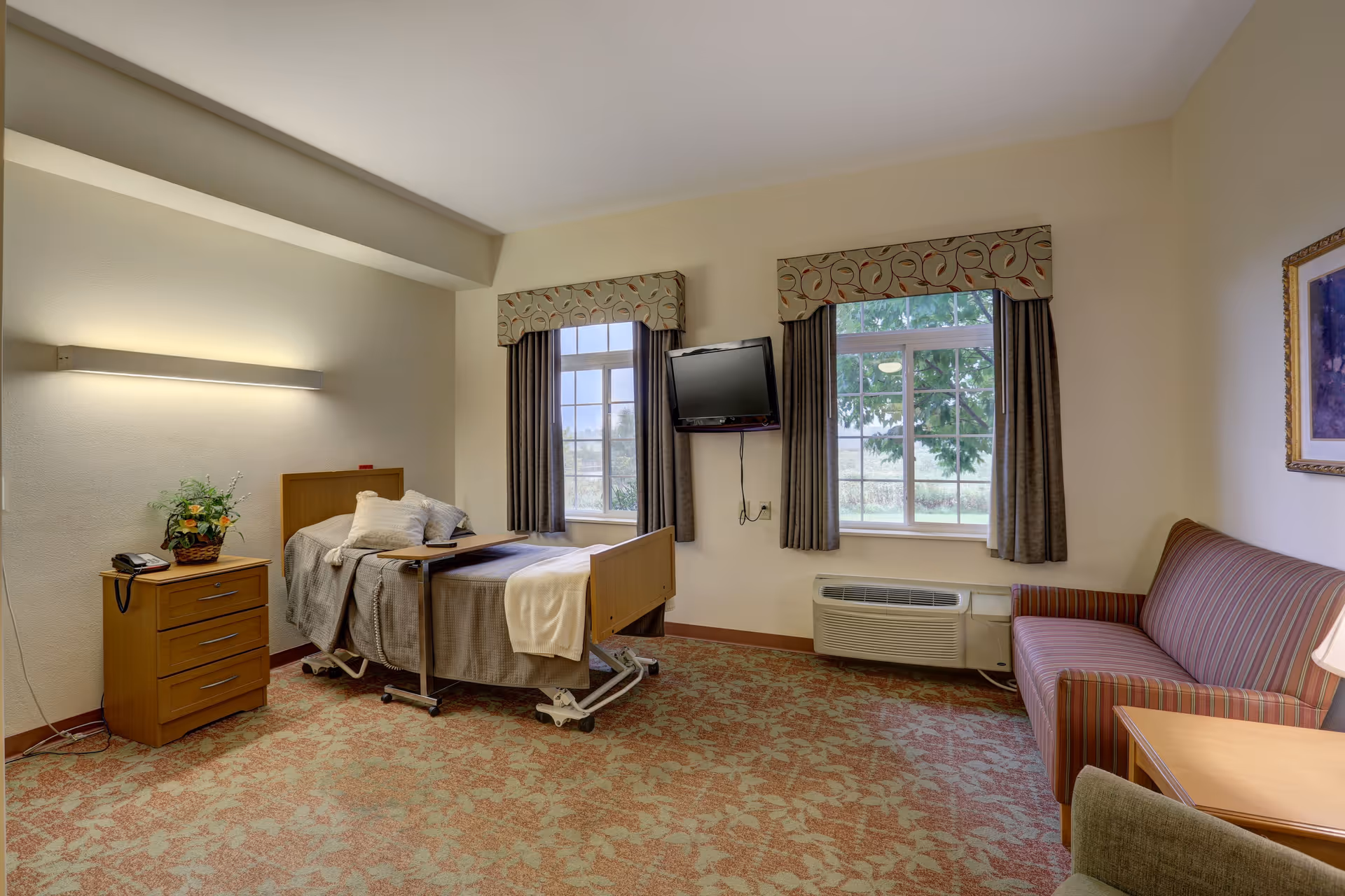A well-lit senior living facility room with a hospital-style bed covered with blankets and pillows, a wooden nightstand with a telephone and a flower arrangement, two windows with patterned valances and curtains, a wall-mounted flat-screen TV, a striped sofa, and a framed picture on the wall.