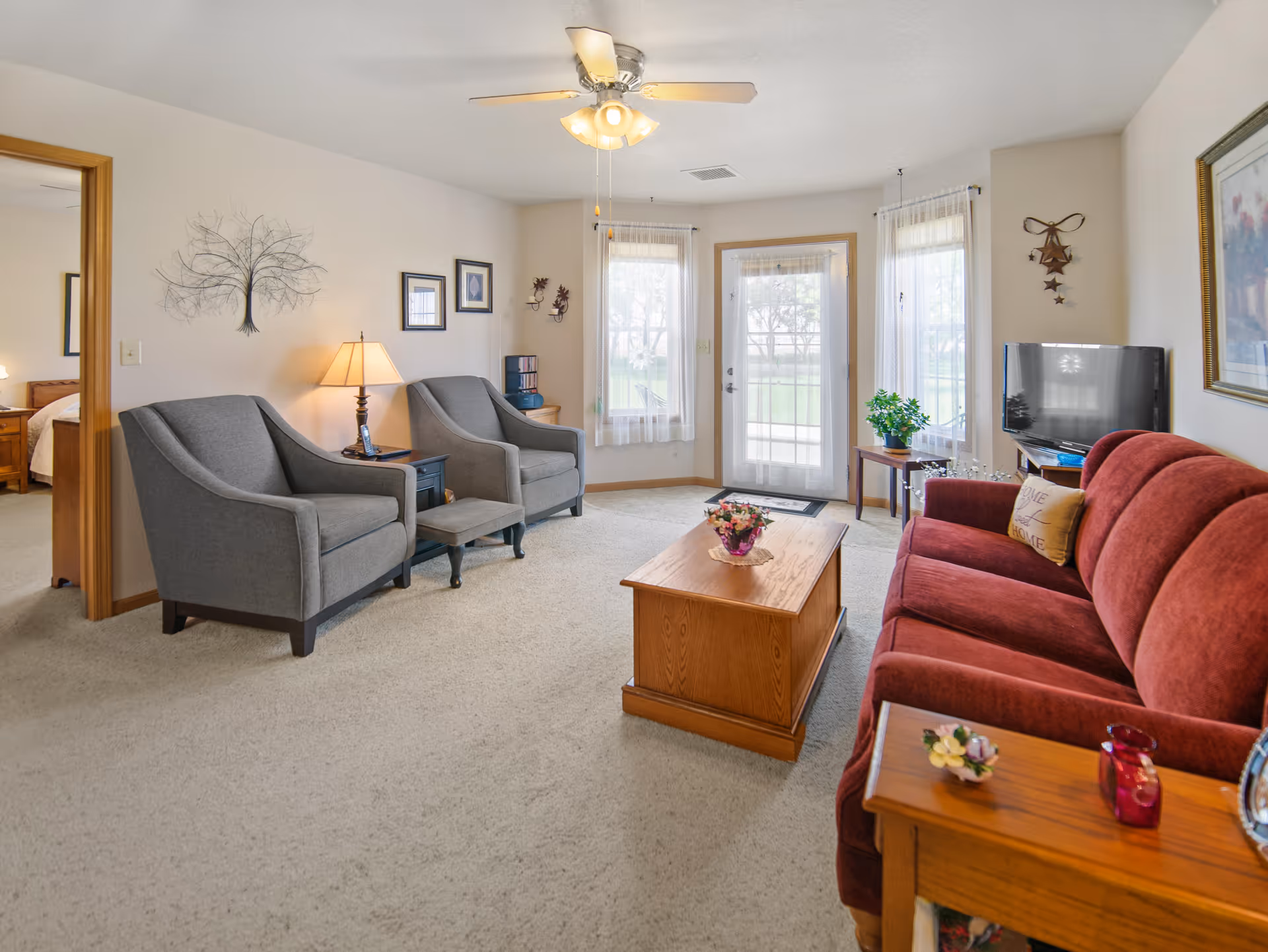 A cozy living room with a red sofa, two gray armchairs, a wooden coffee table with a flower arrangement, and a TV on a stand. The room has beige carpet, light-colored walls with framed artwork, a ceiling fan with lights, and large windows with sheer curtains letting in natural light. A door leads outside to a green lawn. A bedroom is visible through an open doorway on the left.