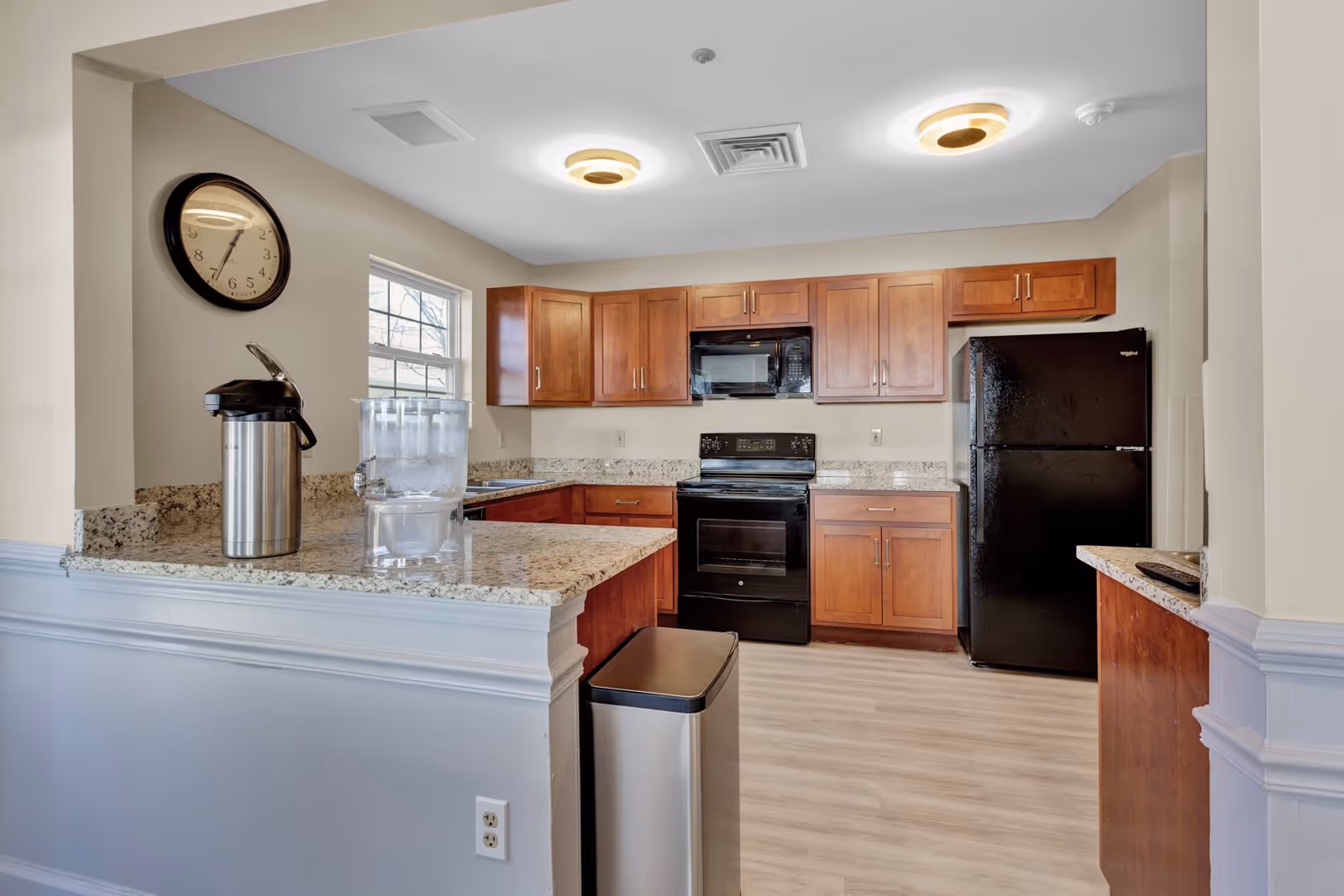 A kitchen area with wooden cabinets, black appliances including a refrigerator, oven, and microwave. There is a granite countertop with a water dispenser and a stainless steel airpot. A wall clock is mounted on the left wall above the countertop. The floor is light-colored wood, and the ceiling has two modern circular light fixtures.