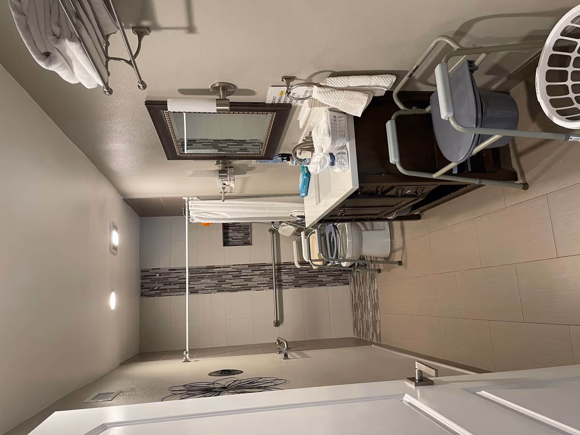 A senior-friendly bathroom with beige tiled floor and walls. It features a walk-in shower with a vertical strip of decorative tiles, a white shower curtain, and grab bars. There is a toilet with a raised seat and armrests, a dark wood vanity with a white countertop and sink, a large framed mirror, and wall-mounted light fixtures. Towels are neatly folded on a metal rack above the vanity, and a commode chair is positioned near the sink. A laundry basket is visible on the floor.
