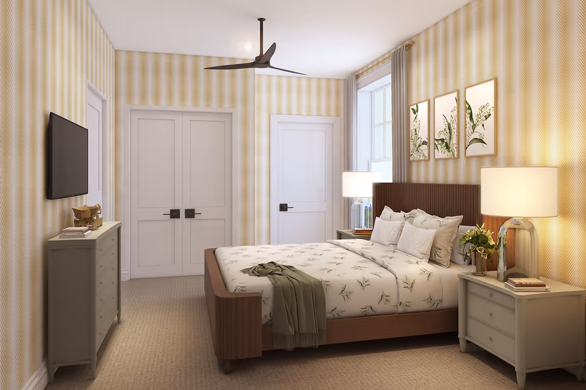 A cozy bedroom with a wooden bed frame and floral bedding. The room features two bedside tables with lamps and a vase of flowers on one side. There is a dresser with books and a decorative item, a wall-mounted TV, and three botanical prints above the bed. The walls have a yellow patterned wallpaper, and there is a ceiling fan overhead. Two white doors and a window with curtains are also visible.