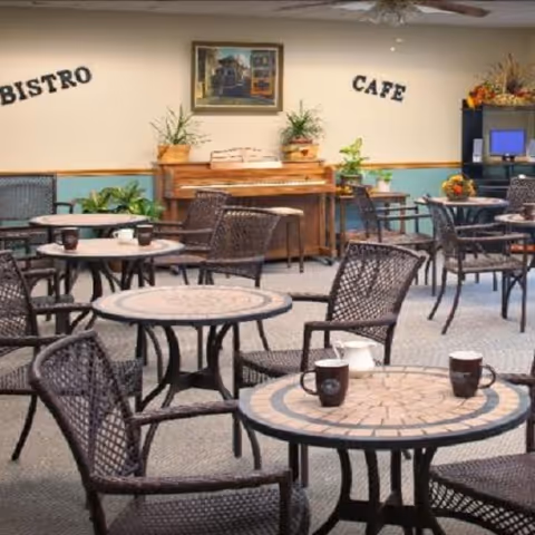 Indoor seating area with round mosaic tables and wicker chairs arranged in a cafe setting. A wooden piano is against the wall with plants on top, and the words 'BISTRO' and 'CAFE' are displayed on the wall above. There is a painting hanging above the piano and a cabinet with a computer monitor and decorative items in the corner.
