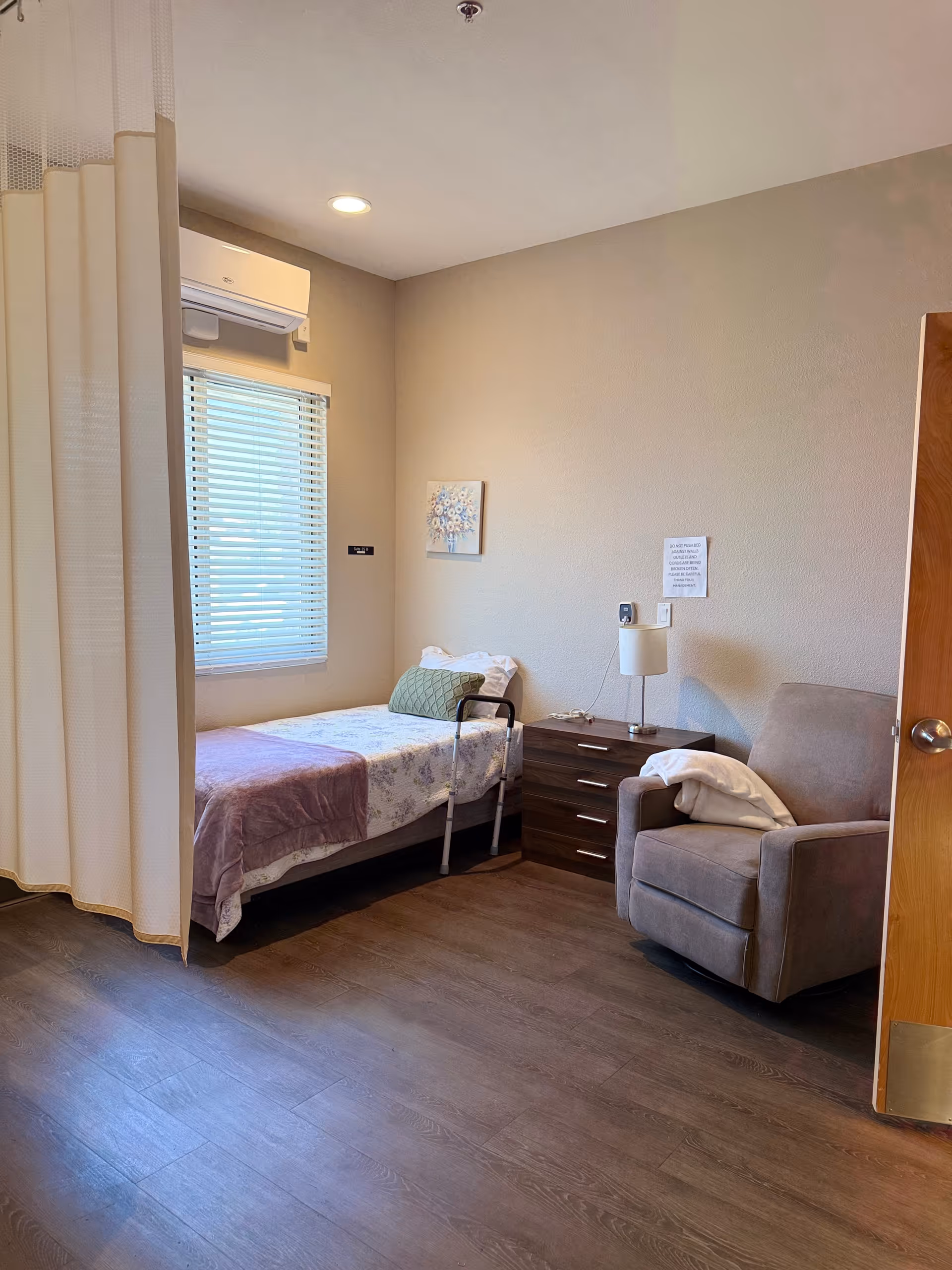 A small memory-care bedroom with a single bed, bedside dresser and lamp, an armchair, and a privacy curtain.