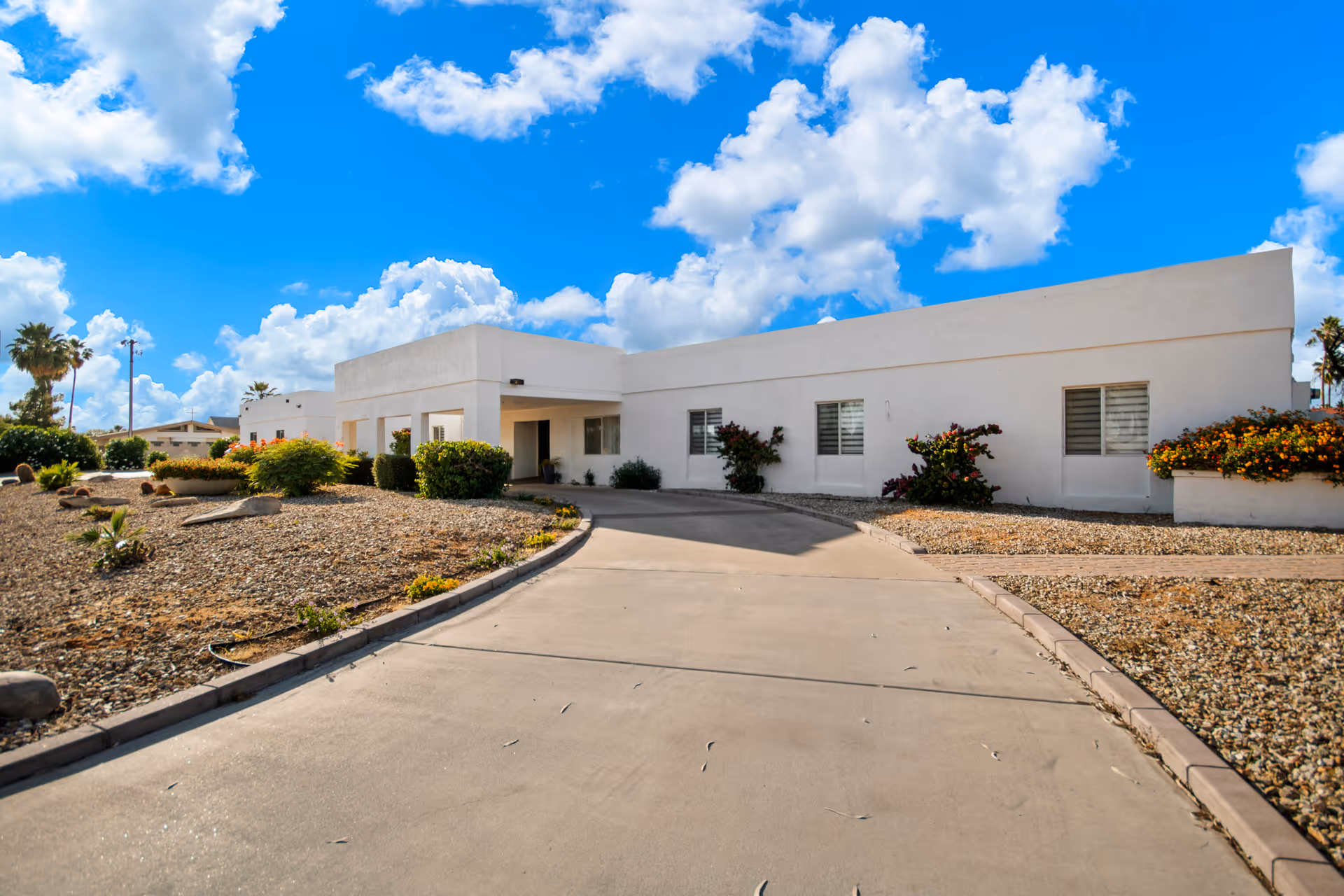 Exterior view of a single-story white building with a flat roof, surrounded by desert landscaping with rocks, bushes, and flowering plants under a bright blue sky with scattered clouds.