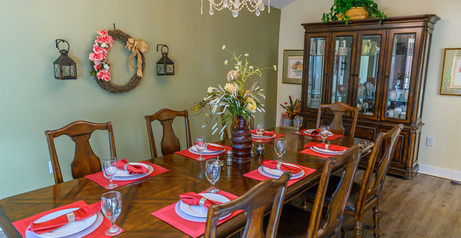 Dining room with a long wooden table set for eight with red placemats and napkins, a floral centerpiece, wooden chairs, and a china cabinet.
