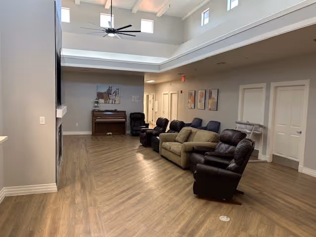 Spacious communal lounge with multiple recliners and sofas arranged along one wall under a high ceiling with skylights and a piano at the far end.