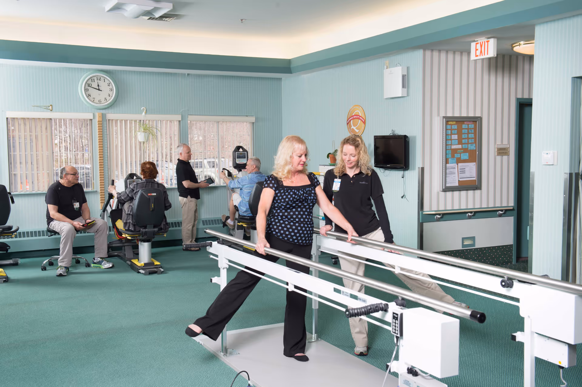 A rehabilitation or physical therapy room in a senior living facility with exercise equipment and parallel bars. A woman is performing a leg exercise on the parallel bars with the assistance of a staff member. In the background, three other seniors are using exercise machines, and a clock on the wall shows the time as 11:55.