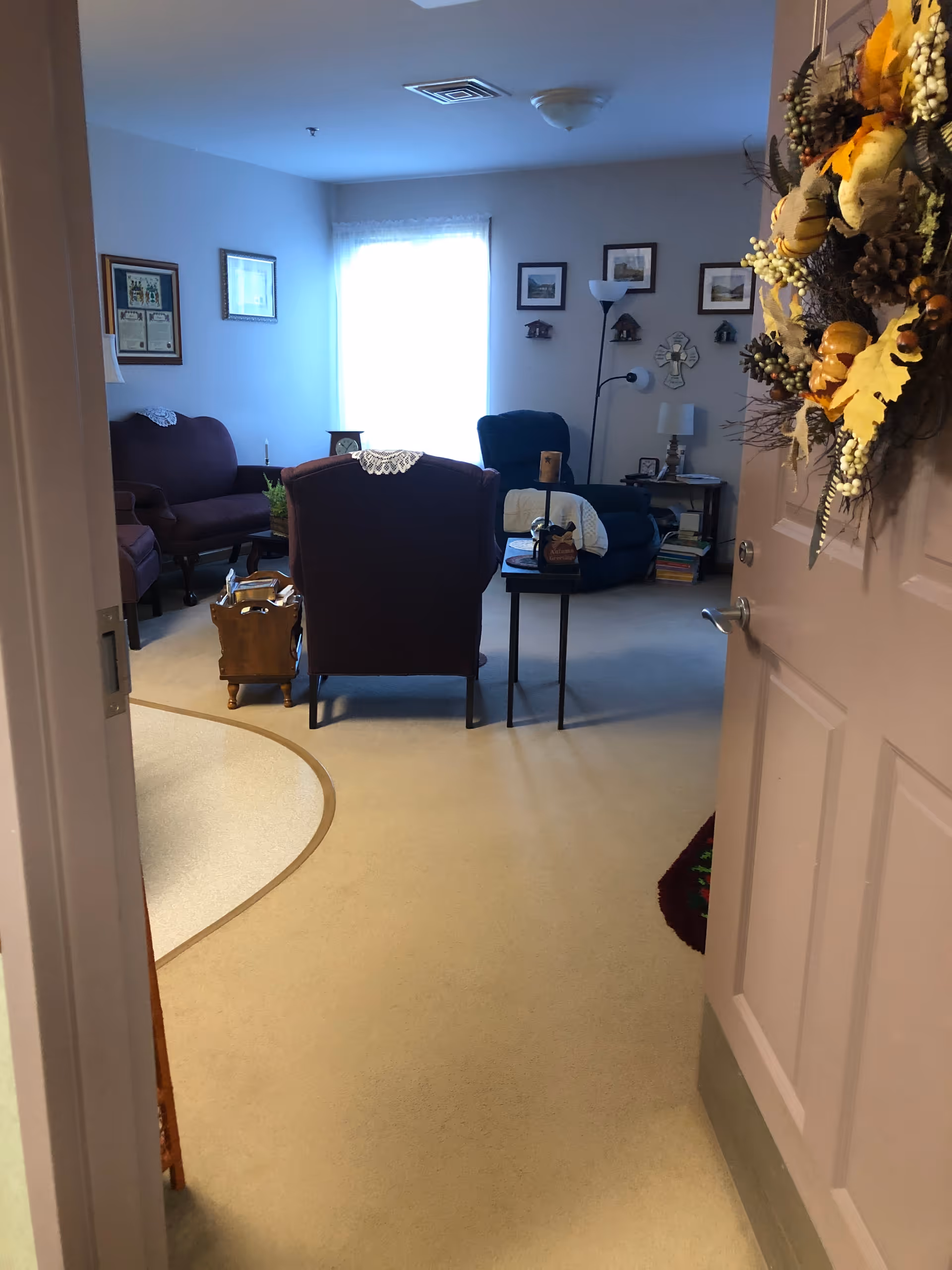 View into a cozy living room area from an open door decorated with a fall-themed wreath. The room has carpeted flooring, a window with sheer curtains letting in natural light, several upholstered chairs, a small table with decorative items, framed pictures on the walls, and a floor lamp.