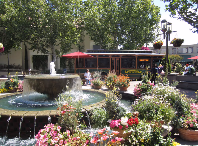 A vibrant outdoor courtyard featuring a large circular water fountain surrounded by colorful flowers and plants. In the background, there is a trolley labeled 'The Fuze Ride' with people sitting and standing nearby. Trees provide shade, and hanging flower baskets adorn lamp posts.