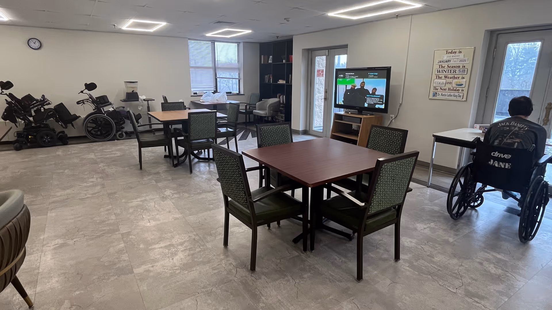 A spacious common room with several tables and chairs arranged for group seating. There are multiple wheelchairs and mobility scooters lined up against the wall on the left. A television is mounted on a stand near the windows and a person in a wheelchair is seated near the right side of the room facing the windows. The room has large windows letting in natural light and a clock on the wall.
