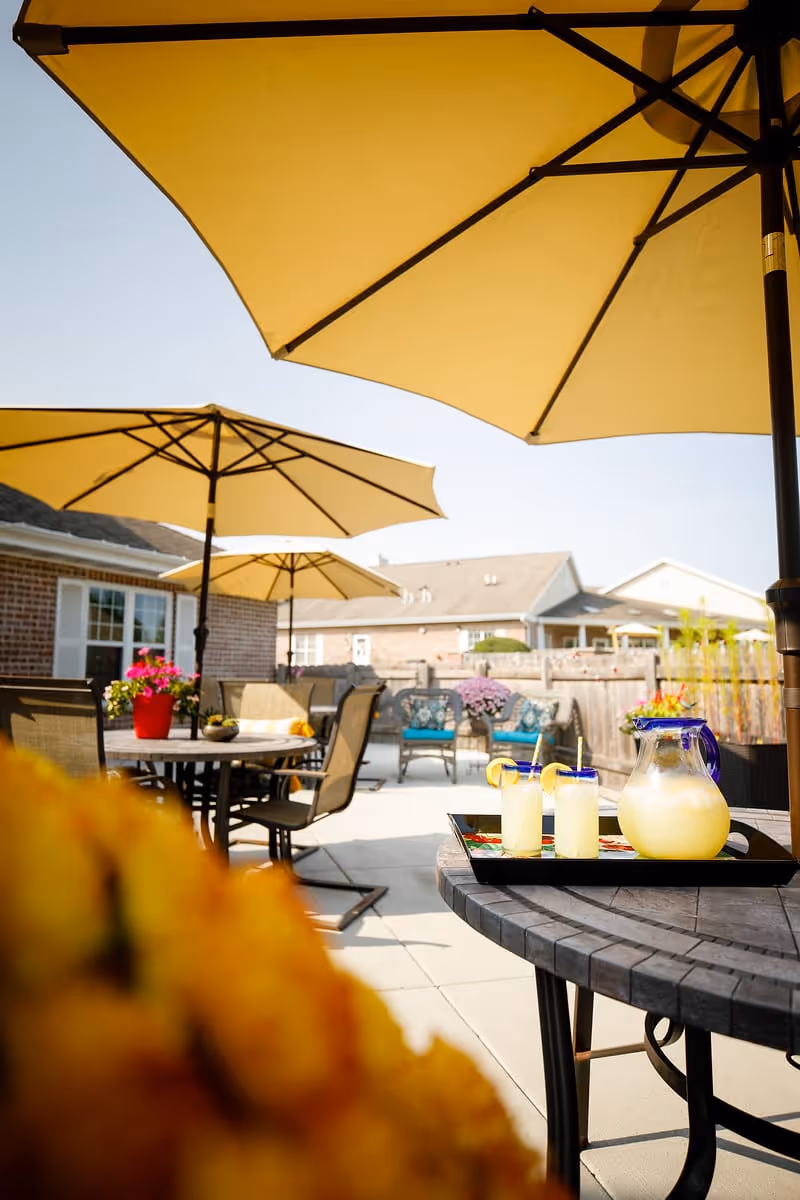 Outdoor patio area with several tables and chairs under large yellow umbrellas. A tray with a pitcher of lemonade and two glasses garnished with lemon slices is on a round table in the foreground. There are potted flowers and a wooden fence surrounding the area, with residential-style buildings in the background under a clear sky.