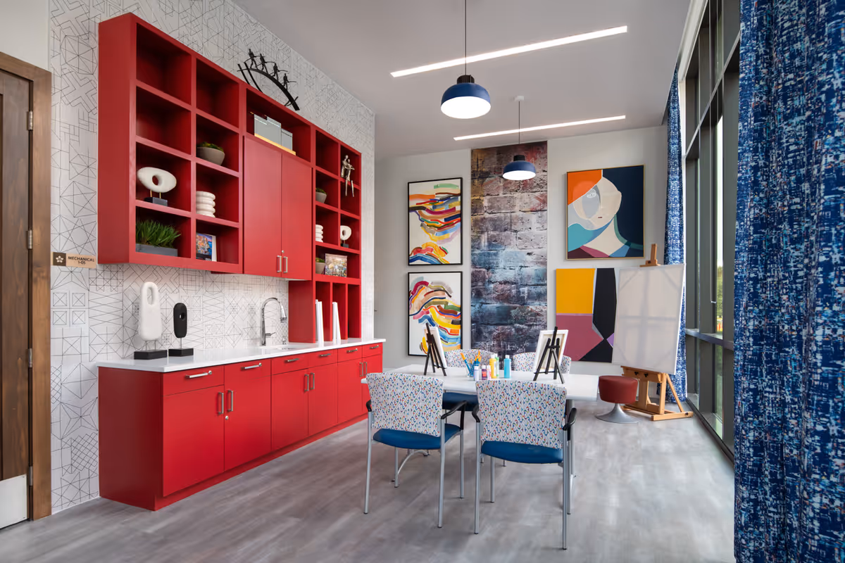 A bright and modern art activity room with a long red cabinet and open shelving on the left wall, decorated with various art pieces and plants. In the center, a white table with four colorful patterned chairs is set up with art supplies, small easels, and paint bottles. The back wall features vibrant abstract paintings and a large window with blue patterned curtains on the right side, allowing natural light to fill the room.